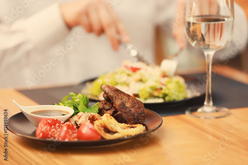 Plate with tasty meat, fried vegetables and sauce on table in restaurant with client on background