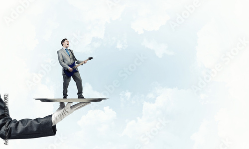 Businessman on metal tray playing electric guitar against blue sky background © adam121