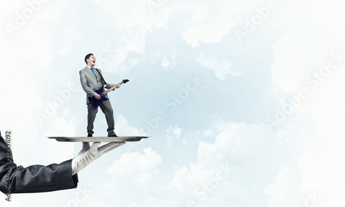 Businessman on metal tray playing electric guitar against blue sky background