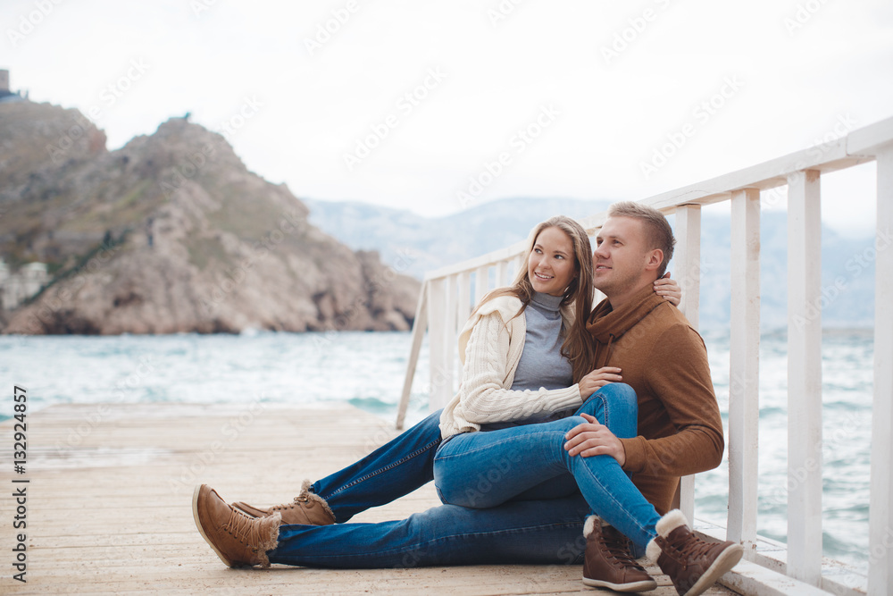 Young couple blond guy with short hair in a brown sweater and blue jeans and a blonde girl with straight long hair,spend time together,sitting,embraced on a white wooden pier near the sea in autumn