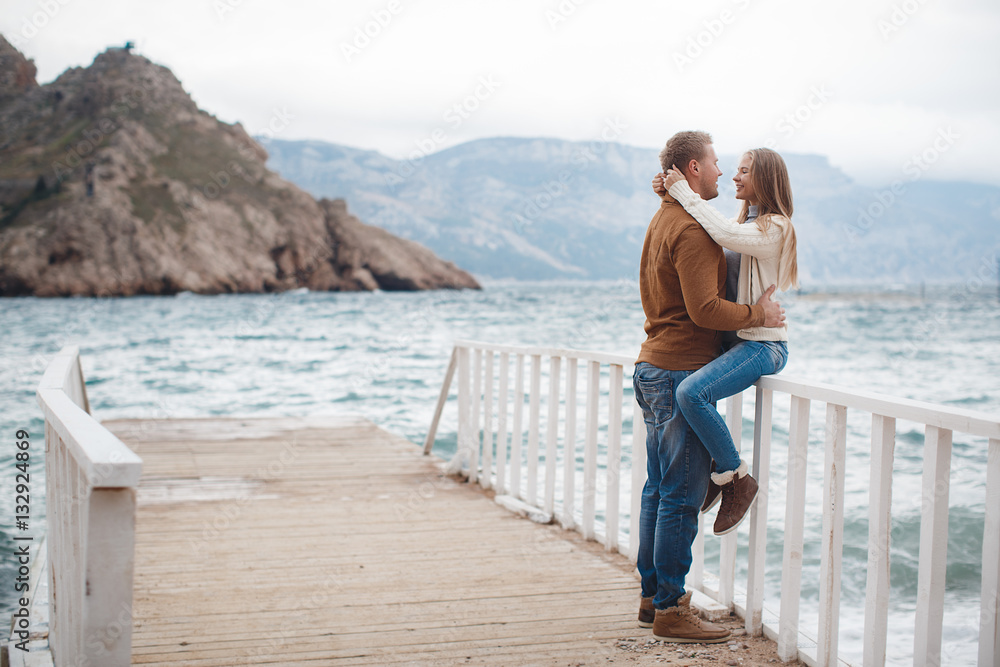 Young couple blond guy with short hair in a brown sweater and blue jeans and a blonde girl with straight long hair,spend time together,standing arm in arm on a white wooden pier near the sea in autumn Young couple blond guy with short hair in a brown sweater and blue jeans and a blonde girl with straight long hair,spend time together,standing arm in arm on a white wooden pier near the sea in autumn