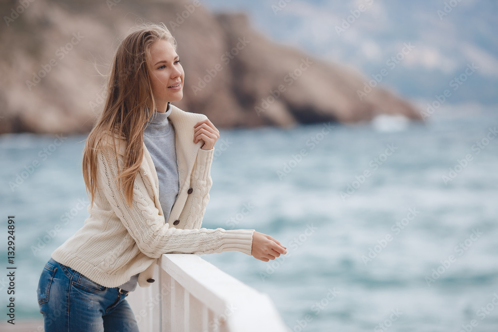 Young woman with long straight blond hair and gray eyes, dressed in a white knitted jacket, a gray turtleneck and blue jeans, spending time alone, standing on white wooden wharf near the blue sea 