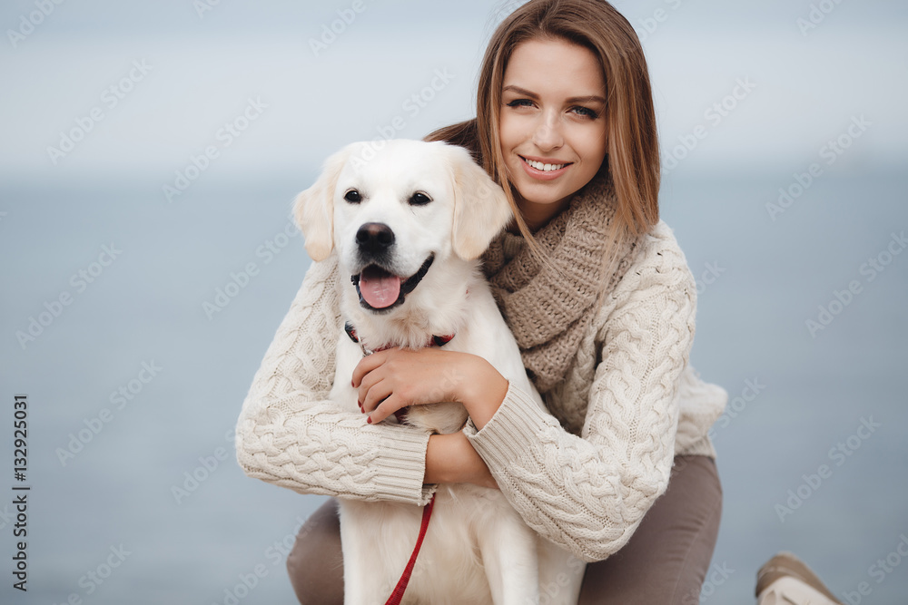 Beautiful young woman with long straight hair, light red,dressed in a white  knitted sweater with a big beige collar,spending time sitting on the beach  with a friend,pet,dog,breed Golden Retriever Stock Photo |