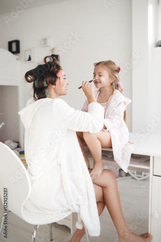 A little girl with long blond hair and her young mother,brunette,dressed in dressing gowns,using cosmetic brushes mother and daughter are doing makeup together,hair wound on large curlers pink