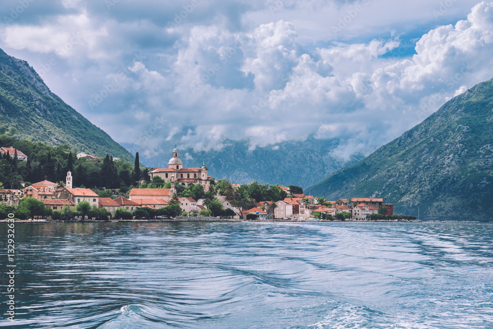 Traditional montenegrin village on Kotor bay by cloudy day in Montenegro. View to red roofs of stone serbian houses, clouds and mountains from water of Boka Kotorska.