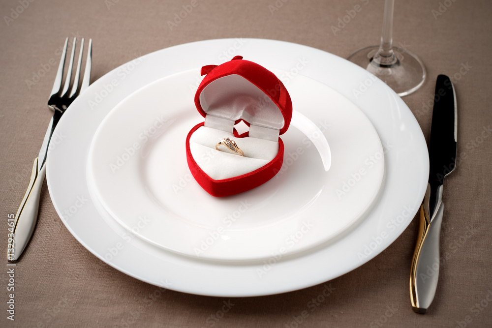 Golden ring in red case on white plate with fork, knife and glass. Golden ring in red case on white plate with fork, knife and glass.