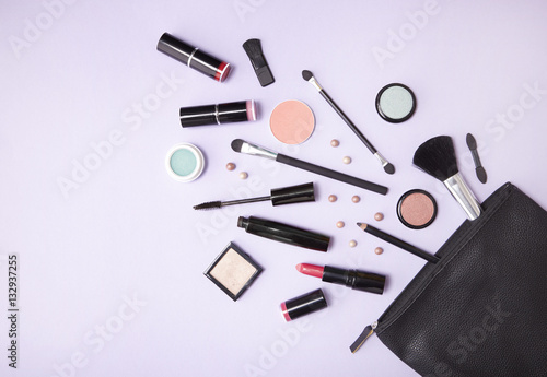 A black leather make up bag with cosmetic beauty products spilling out on to a pastel purple background, with blank space at side