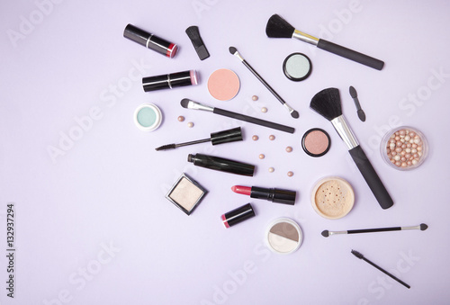 A collection of make up and cosmetic beauty products strewn over a pastel purple background with blank space at side