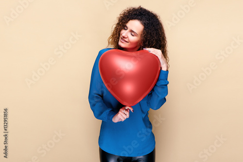Beautiful young girl with curly hair and blue sweater holding a red heart air balloon. The concept of Valentine's Day © khmelev