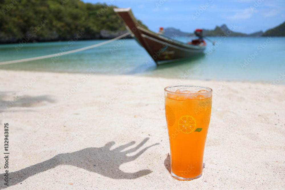 Glass of orange juice and shadow of hand on the beach with views of the archipelago Islands and long-tail boats. 