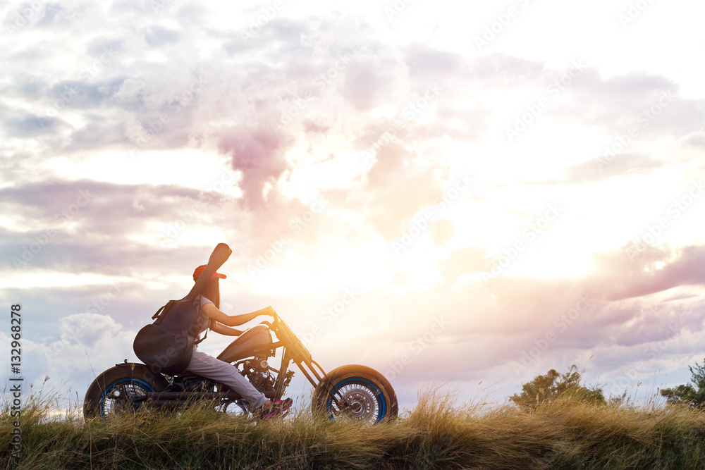 Fototapeta premium Guitarist woman riding a motorcycle on the countryside road, sunset background