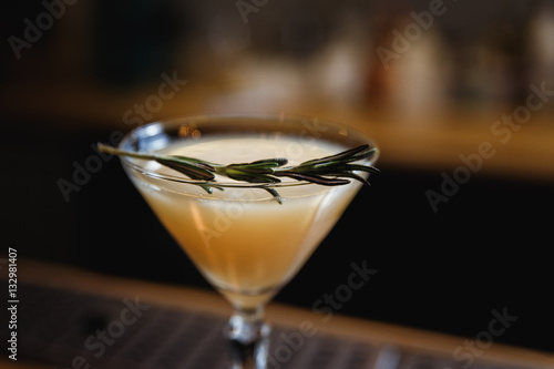 A drink on the bar stand in a cocktail (martini) glass with rosemary on top. Small depth of field, selective focus, film grain effect.