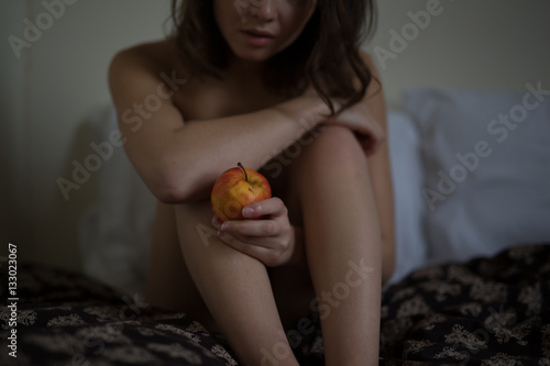Naked female hipster plays with a red apple on bed and thinks de photo