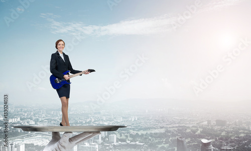 Attractive businesswoman on metal tray playing electric guitar against cityscape background