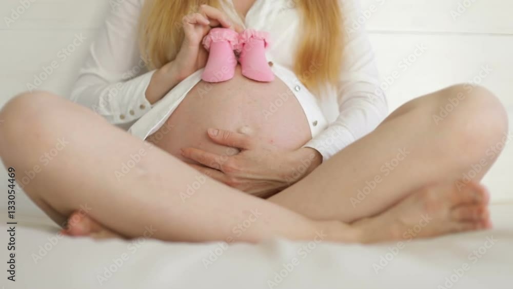 Waiting for baby with baby's booties. Young pregnant woman sits and stroking belly. She holds the pink baby's bootees. Waiting for baby with baby's booties. Young pregnant woman sits and stroking belly. She holds the pink baby's bootees.