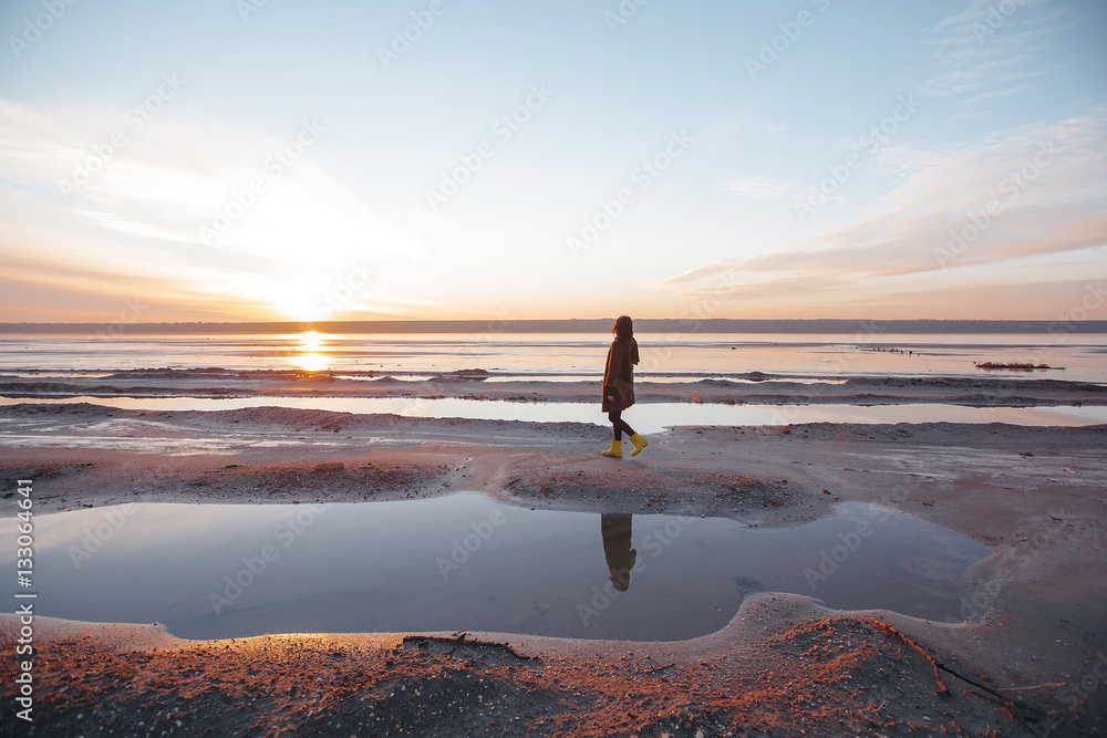 Beautiful girl is standing on the sea in a Sunbeam, yellow boots, long black coat, sand underfoot