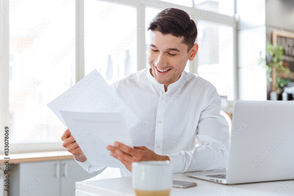 Happy young man holding documents while using laptop Happy young man holding documents while using laptop