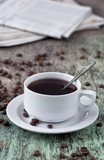 A cup of black coffee on the wooden table with coffee beans are scattered. Black coffee hot drink Grains Newspaper Wooden background White cup Breakfast