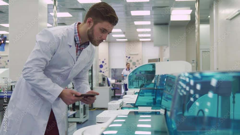 Attractive caucasian man in white coat flipping on the tablet near laboratory machine. Young male scientist coming close to the lab equipment. Handsome bearded guy looking at the centrifugal machine Attractive caucasian man in white coat flipping on the tablet near laboratory machine. Young male scientist coming close to the lab equipment. Handsome bearded guy looking at the centrifugal machine