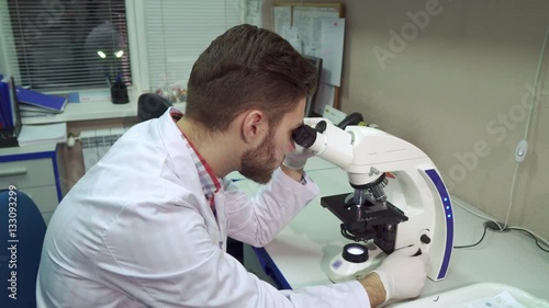 Handsome caucasian man moving stage clipse on the scope. Young male scientist working with microscope at the laboratory. Attractive bearded guy using optical lab tool photo