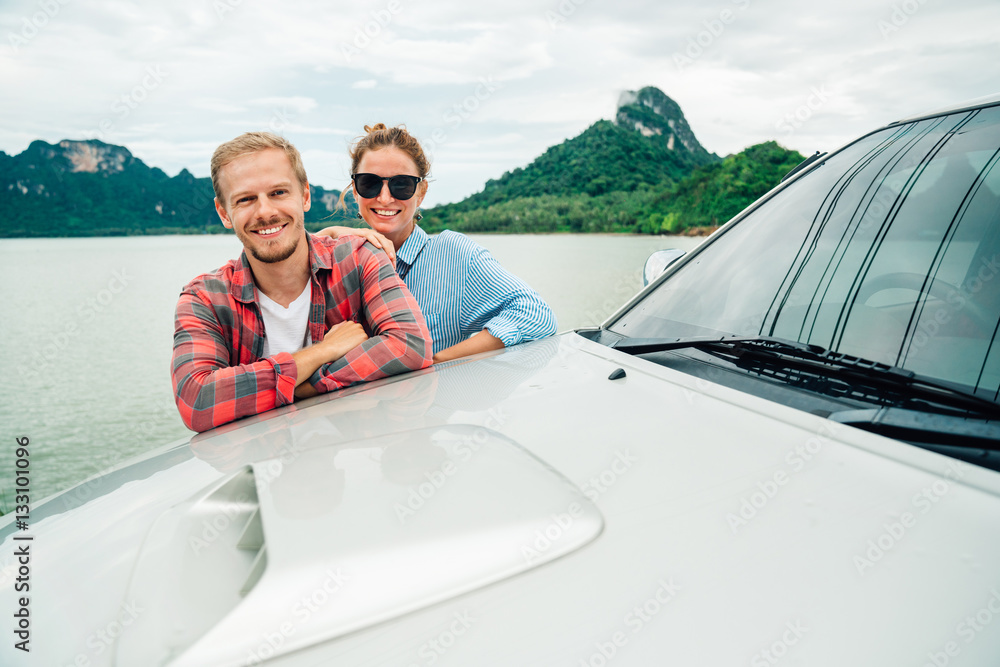Naklejka premium young couple traveling by car. portrait of a man and woman embracing and looking at camera on the background of mountain scenery of the lake and sky. romantic vacation and travel.
