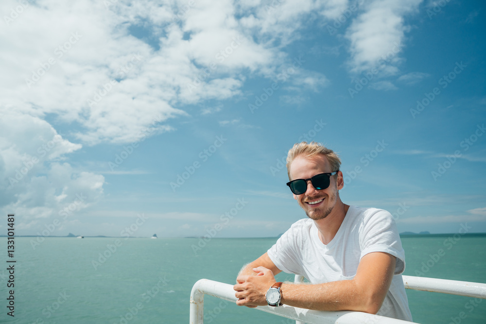 Portrait of a young successful man on the background of sky and sea landscape..Blonde with glasses in white shirt leaning on the railing of the ship.