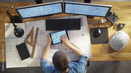 Top View of architectural Engineer Draws on His Blueprints, Compares with Tablet Computer with Green Screen, Using Desktop Computer Also. His Desk is Full of Useful Objects and Evening Sun. 