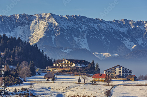 Sunny winter scenery with majestic snowy Piatra Craiului mountains, chalet or lodging buildings in Cheile Gradistei Fundata touristic resort, Romania. Idyllic, quiet holiday retreat destinations. photo