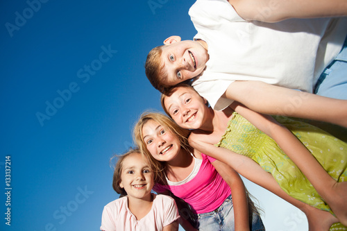 four happy beautiful children looking at camera from top in the sunny summer day and blue sky. looking at camera with funny face and toothy smile. view from below.
