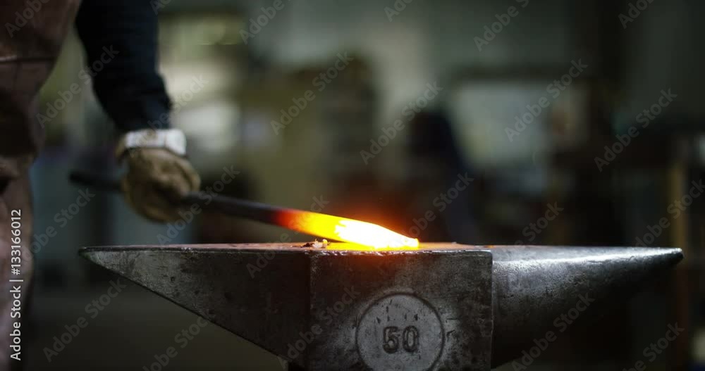 In industry a blacksmith,working hot iron tapping it with a hammer on anvil,and causes hot sparks.Behind the blacksmith a worker using the welding machine.Concept:industry,handicrafts,iron and steel