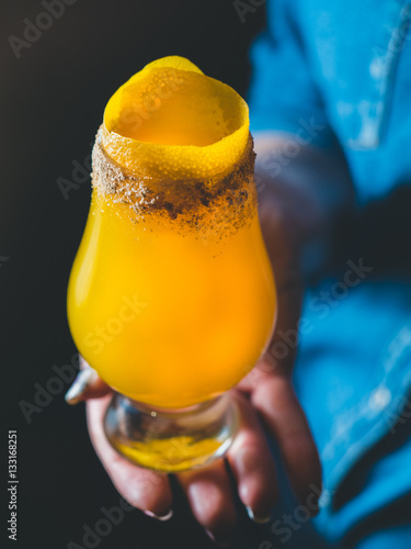 Yellow mango cocktail with lemon zest and sugar crusta in a tulip glass, female hand / bartender holding a glass with the cocktail, blue jeans shirt background. Cocktail close-up, vertical orientation photo