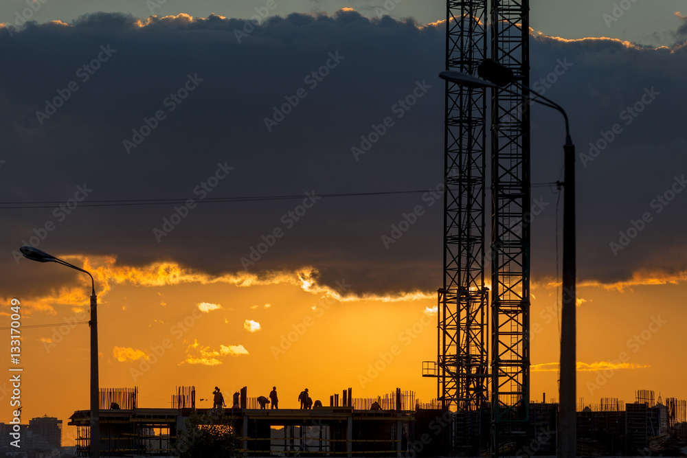 Construction workers silhouettes, cranes and building construction site against sunset sky.