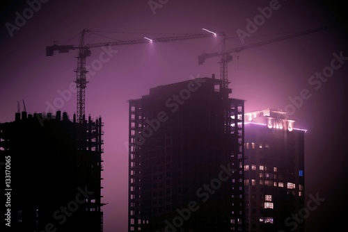 Misty cityscape at night time. Cranes over houses under construction.