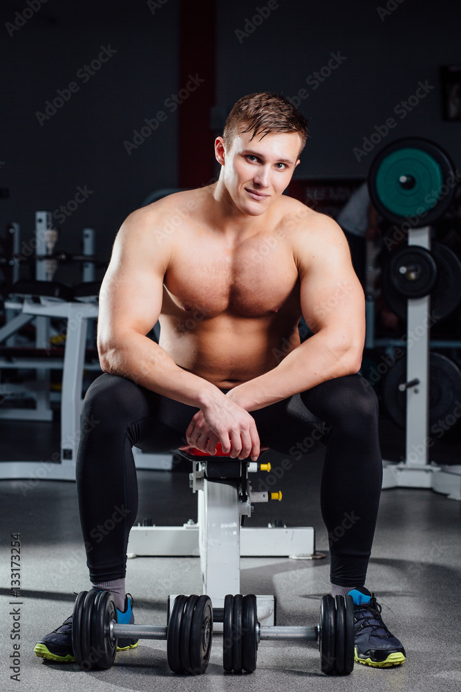 professional bodybuilder sitting on the bench, resting between exercises with dumbbells at gym. Big