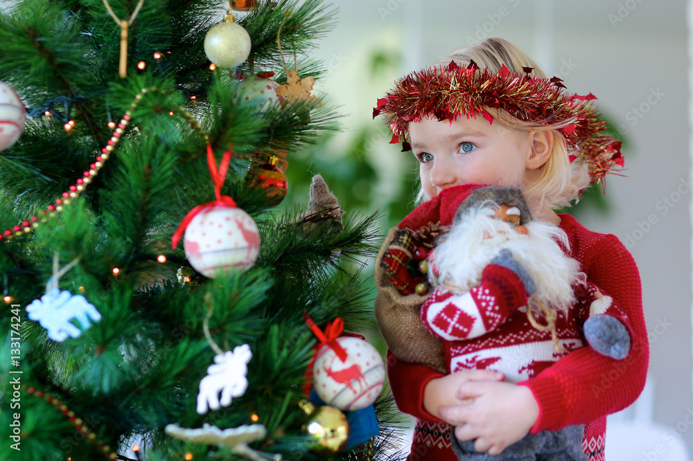 Little girl playing with Santa toy under decorated christmas tree. Cute kid in Xmas mood preparing home for family celebration. Little girl playing with Santa toy under decorated christmas tree. Cute kid in Xmas mood preparing home for family celebration.