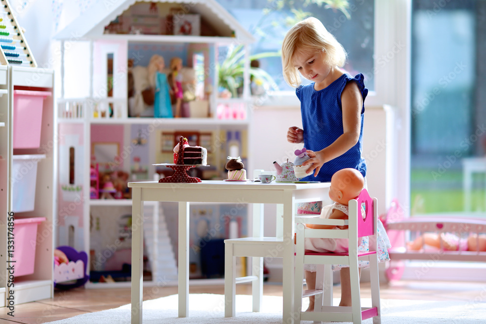 Cute preschooler girl having birthday tea party with her doll. Little child plays in sunny room at home or kindergarten. Kids using toy dishes and cakes. Cute preschooler girl having birthday tea party with her doll. Little child plays in sunny room at home or kindergarten. Kids using toy dishes and cakes.