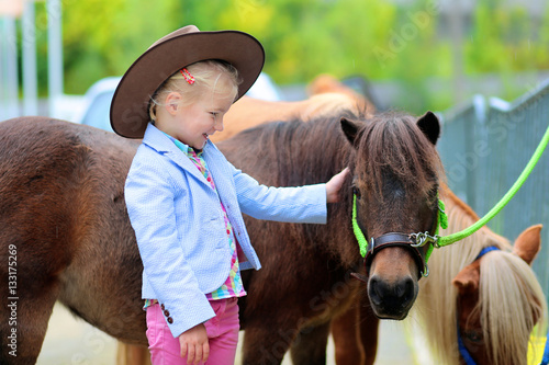 Lovely cowgirl caressing little pony horse in the farm. Pretty preschooler girl wearing cowboy hat playing with animals outdoors on sunny day.
