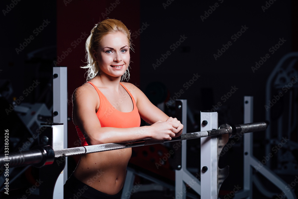 Blonde fitness girl with perfect shape body rests after sport workout in gym leaning on barbell Blonde fitness girl with perfect shape body rests after sport workout in gym leaning on barbell