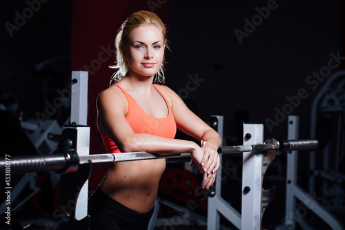 Blonde fitness girl with perfect shape body rests after sport workout in gym leaning on barbell