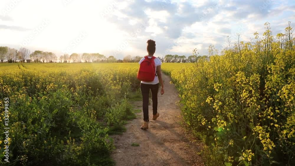 Beautiful happy mixed race African American girl teenager female young woman hiking with red backpack in field of rape seed yellow flowers Beautiful happy mixed race African American girl teenager female young woman hiking with red backpack in field of rape seed yellow flowers