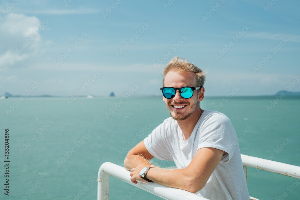 Portrait of a young attractive guy in a white dress and sunglasses on the background of the celestial landscape. Blonde with glasses in white shirt leaning on the railing of the ship. Portrait of a young attractive guy in a white dress and sunglasses on the background of the celestial landscape. Blonde with glasses in white shirt leaning on the railing of the ship.