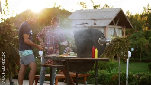 Two Caucasian males making hamburgers with fried meat and buns near barbecue grill in slowmotion