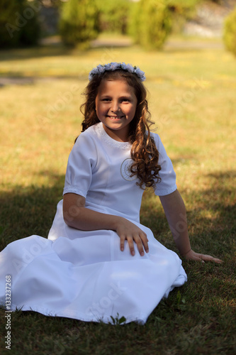 Śliczna dziewczynka w białej sukni siedzi na trawie.