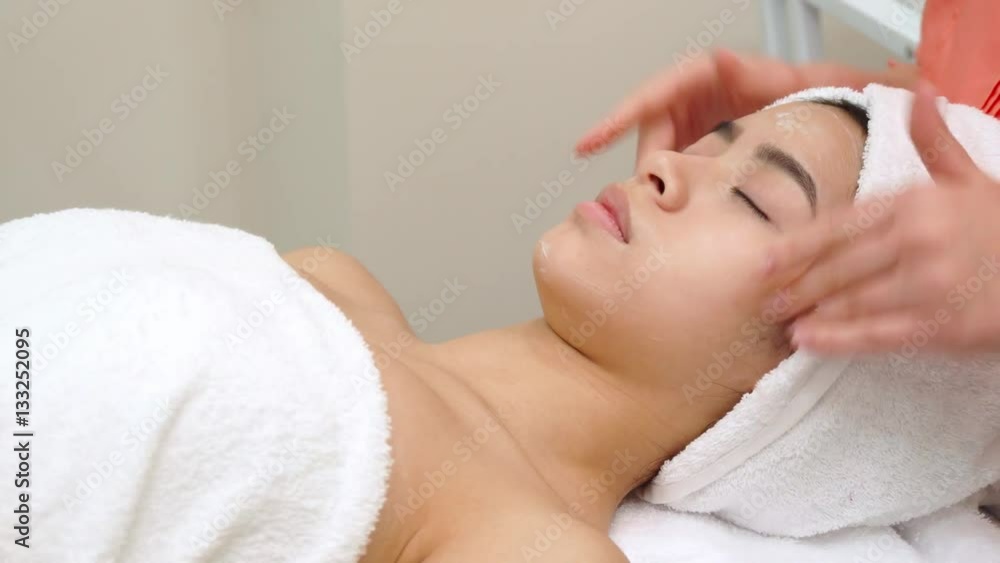 Masseur massaging girl's face at the beauty salon. Side view of female hands stroking cheeks and forehead of pretty asian client. Attractive brunette woman lying on massage chair Masseur massaging girl's face at the beauty salon. Side view of female hands stroking cheeks and forehead of pretty asian client. Attractive brunette woman lying on massage chair