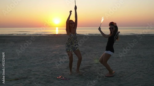 Two euphoric young women partying with firework candles on the beach at sunrise