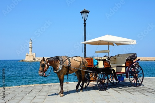 Horse drawn carriage on the quayside with the Venetian lighthouse at the harbour entrance to the rear, Chania, Crete.