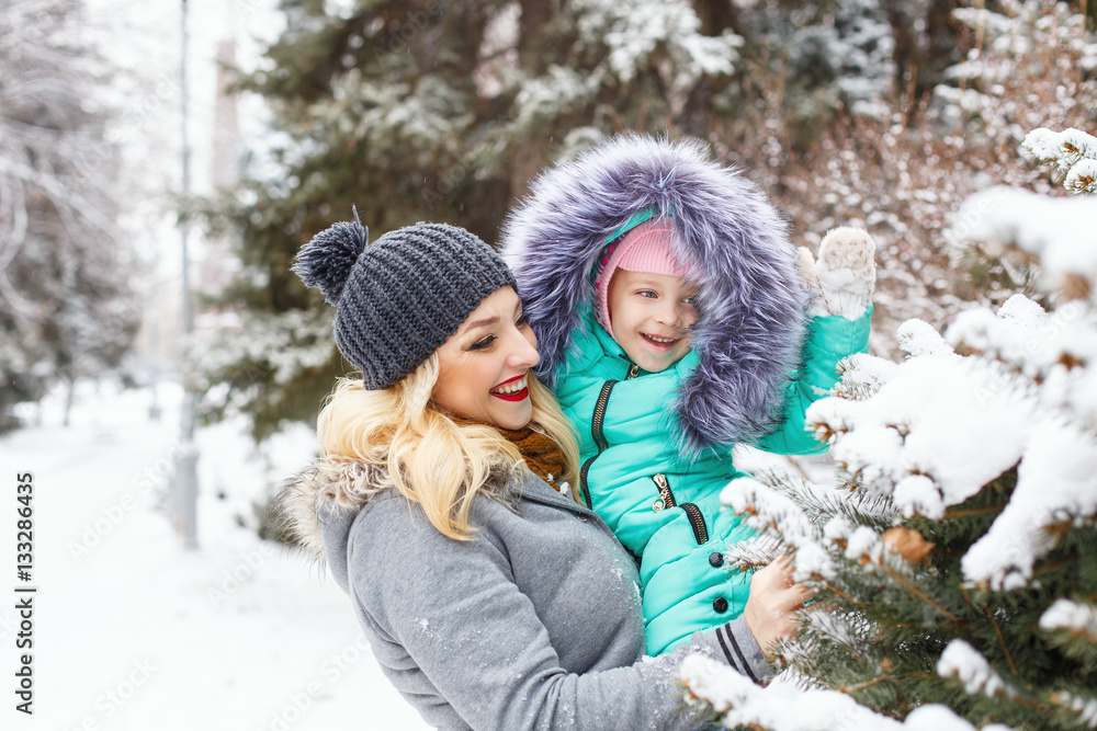 Mother holds daughter on hands. They stand next to the Christmas tree in snow. Family time. Winter walk in the park.
