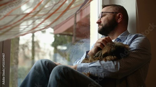 Sad man looking out the window with a dog on hands

