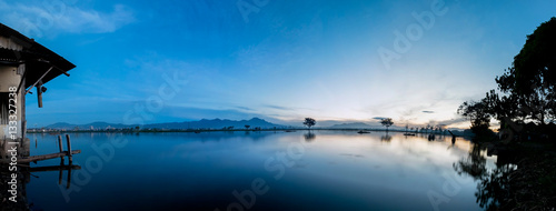 Panorama photo of very vast, broad, large, spacious pond, stretched into the horizon. Behind it is a line of hills and mountains, silhouette of tree, house and beautiful blue sky. This photo captured