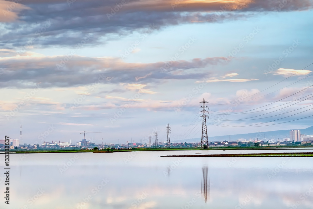 Transmission tower, electricity pylon or a power pylon, standing majestically lined up to the horizon, in front of large lake or pond, also with beautiful cloud and city in the background, captured at